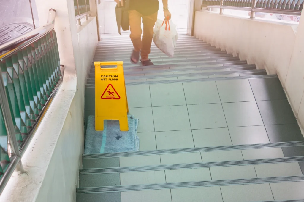 Person walking down tiled stairs near a "Caution Wet Floor" sign placed on a mat at the top of the steps, indicating a potential slip hazard.