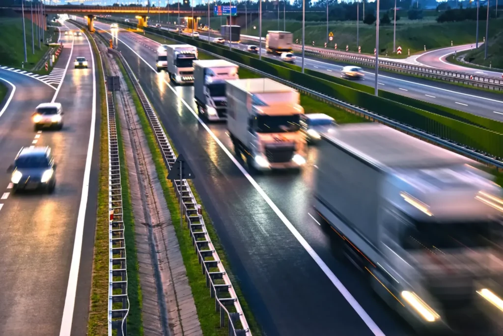 Multiple semi-trucks and vehicles driving along Interstate 10 at dusk, with wet roads reflecting headlights and traffic flowing in both directions.