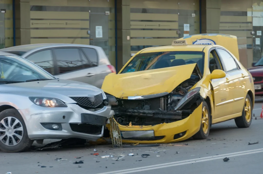 Front-end collision between a silver sedan and a yellow taxi on a city street, with visible damage and scattered debris.