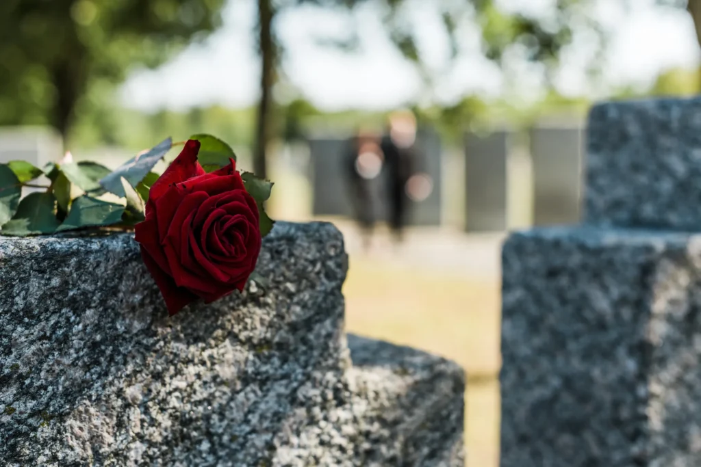 Single red rose resting on a granite gravestone in a cemetery, with blurred mourners in the background.