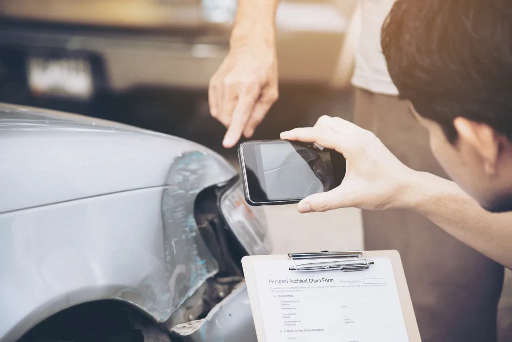 Man photographing damage on a silver car while another person points, with a clipboard displaying a personal accident claim form in the foreground.