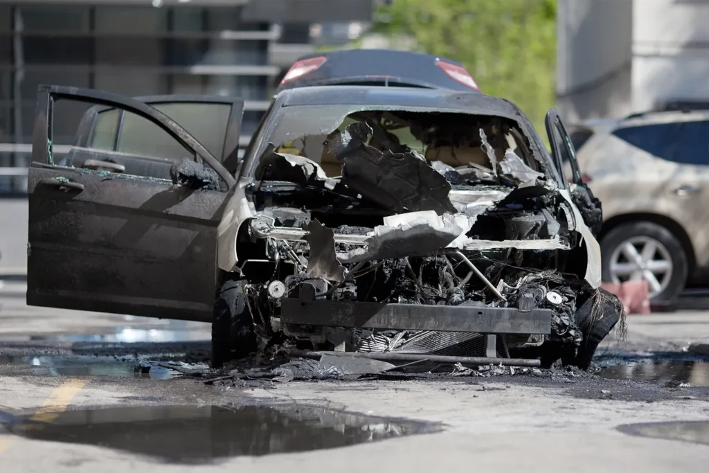 Burned and destroyed car with its front completely melted and engine exposed, parked in a city lot after a severe fire.